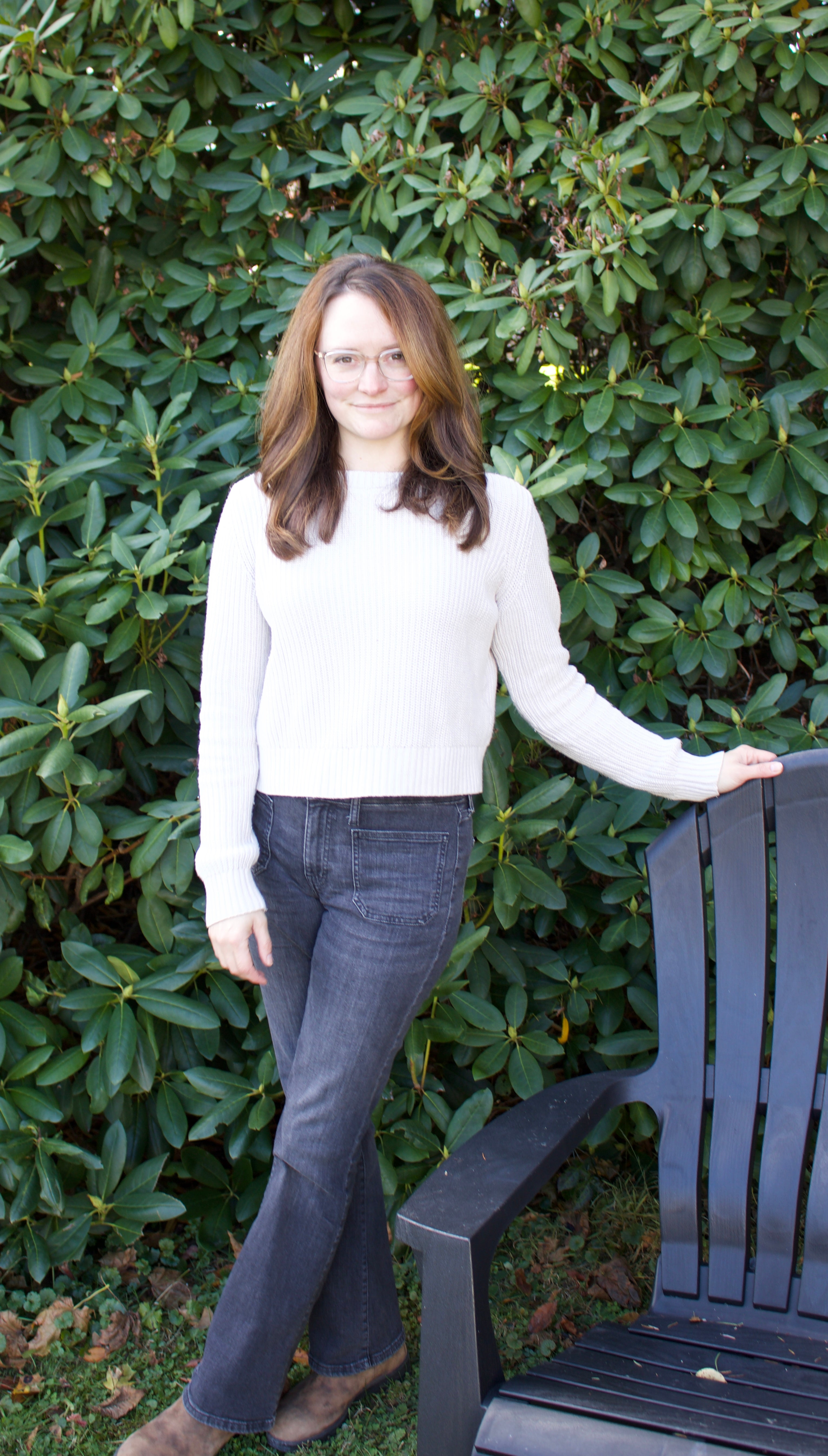 Woman standing next to a chair against a green bush background